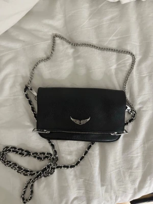 Svart axelväska Zadig & Voltaire - Andvänd få gånger, nytt skick. Nypris: 2900kr. PRIS KAN DISKUTERAS! Snygg svart axelväska i läder från Zadig & Voltaire med silverfärgad kedjerem och ikonisk vingdetalj på framsidan.  Liten och smidig modell med plats för det viktigaste.