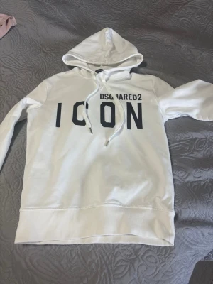 Vit hoodie DSQUARED2 ICON - Snygg vit hoodie från DSQUARED2 med stor svart 'ICON'-text på bröstet och justerbar huva med dragsko. Klassisk passform med ribbade muddar och nederkant. Perfekt för dig som gillar streetwear och vill sticka ut med ett trendigt plagg.