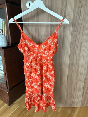 Röd blommig klänning från BikBok - Supersöt röd klänning från BikBok med vita och rosa blommor, smala axelband och volang nertill. Perfekt för varma dagar och har en figurnära passform med elastisk rygg för extra komfort.