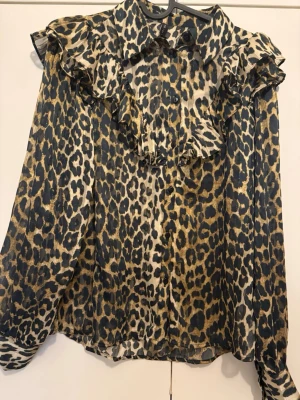 Leopardmönstrad blus med volang - Snygg blus med leopardmönster i svart, beige och guldiga toner. Blusen har långa ärmar, knäppning framtill och volangdetaljer vid axlar och bröst. Tunt, lätt och transparent material som ger en trendig look.