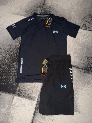 Under Armour träningsset t-shirt & shorts - Snyggt träningsset från Under Armour med t-shirt och shorts. Finns i svart och ljusgrått. Setet har färgglada loggor och detaljer, samt elastisk midja på shortsen. Perfekt för gymmet eller löpning. Materialet är lätt och andas.