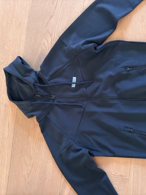 Svart hoodie från djurgården  - Svart hoodie med dragkedja från djurgården . Modellen har huva med dragsko, två fickor med dragkedja och en liten logotyp på bröstet. Perfekt för en chill och sportig stil. Materialet känns mjukt och slitstarkt.