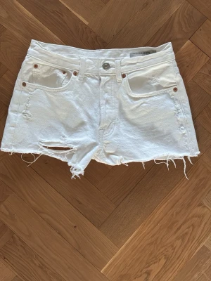 Vita denimshorts med fransar - Säljer ett par vita denimshorts från Alisanne med råa, fransiga kanter och slitna detaljer. Klassisk femficksmodell med knapp och dragkedja framtill. Perfekta för varma dagar och en avslappnad look.