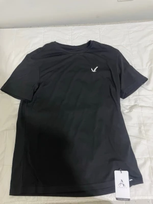 Svart t-shirt från Asrv - Stilren svart t-shirt från Asrv med en vit broderad logga på bröstet. Klassisk rund halsringning och korta ärmar. Tillverkad i mjuk bomull som känns skön mot huden. Perfekt för en clean och enkel look.