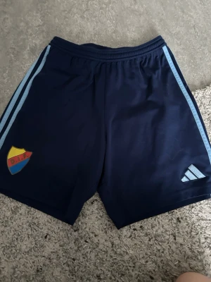 Blå Djurgården shorts från Adidas - Mörkblå sportshorts från Adidas med ljusblå ränder längs sidorna och Djurgårdens IF klubbmärke på ena benet. Perfekta för träning eller match. Elastisk midja för skön passform.