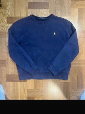 Mörkblå crewneck från Polo Ralph Lauren - Säljer en mörkblå crewneck från Polo Ralph Lauren med klassisk gul logga broderad på bröstet. Tröjan har rund halsringning, ribbade muddar och är tillverkad i mjukt bomullsmaterial. Perfekt för dig som gillar stilrena och tidlösa plagg.