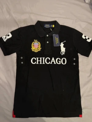 Svart pikétröja Polo Ralph Lauren - Svart pikétröja från Polo Ralph Lauren med stor vit logga på bröstet, broderad gul och röd patch samt texten CHICAGO framtill. Klassisk krage och knappar vid halsen. Siffran 3 på båda ärmarna. Perfekt för en sportig och stilren look.