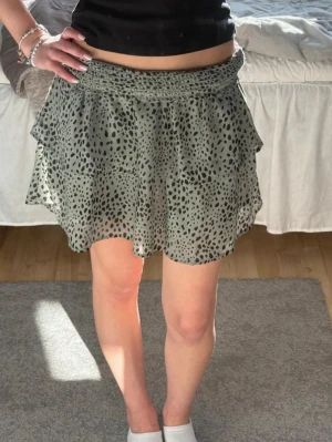Grön kjol - Säljer en grön kortkjol med svart leopardmönster och volangdetaljer. Kjolen har bred smock i midjan för skön passform och är gjord i ett lätt, luftigt material som passar perfekt till varmare dagar.