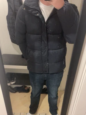Stilren Svart Pufferjacka - Svart pufferjacka från Dressman med dragkedja och huva. Jackan har stora, quiltade sektioner och två sidofickor med dragkedja. Perfekt för kalla dagar och har en klassisk, stilren look som passar till det mesta. Fynda Perfekt vinterjacka! En liten defekt, syns på bild 4, men absolut ingenting som förstör funktionen eller helheten! :)