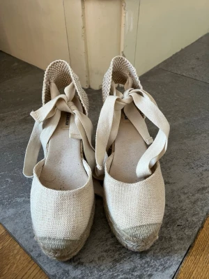 Beiga espadrilles med knytband - Snygga beiga espadrilles med högt flätat repklack och breda knytband runt ankeln. Ovandelen är i grov canvas och bakkappan är stickad för extra detalj. Storlek 38 och ungefär 10cm höga