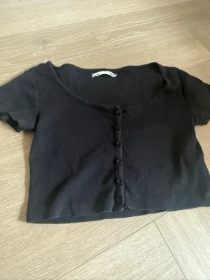 Svart croppad topp med knappar - En svart croppad topp med korta ärmar och knappar framtill. Toppen har en djup rund halsringning och är tillverkad i ett ribbat material som ger en skön och stretchig känsla. Perfekt att matcha med höga jeans eller kjol.