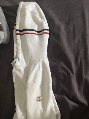 Vit hoodie från Moncler med dragkedja - Säljer en vit hoodie från Moncler med dragkedja, huva med vita snören och Moncler-logga på ärmen. Muddarna har snygga ränder i rött, vitt och blått. Hoodien har fickor framtill och är gjord i mjukt bomullsmaterial. Bra skick!! Storlek S funkar för M