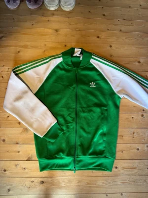 Grön Adidas track jacket - Snygg grön Adidas track jacket med vita ärmar och klassiska tre ränder längs axlarna.