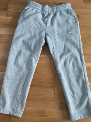 Grå mjukisbyxor från Nike - Snygga grå mjukisbyxor från Nike med elastisk midja och sidofickor. Byxorna har en liten Nike-logga broderad på vänster sida och en bakficka med knapp. Perfekta för chill dagar eller träning.