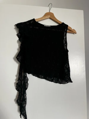 Svart spetsig  topp - Säljer en svart oneshoulder topp i spets med asymmetrisk skärning och volangdetalj längs ena sidan. 