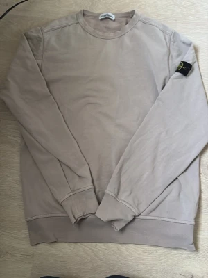 Beige sweatshirt från Stone Island - Snygg beige sweatshirt från Stone Island med klassisk rund hals och ikonisk logotyp-patch på vänster ärm. Tröjan har ribbade muddar och är tillverkad i mjukt bomullsmaterial. Perfekt för en avslappnad och stilren look.