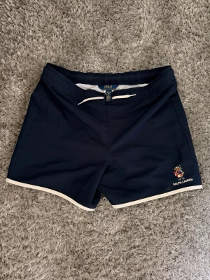 Mörkblå badbyxor från Ralph Lauren - Snygga mörkblå badbyxor från Ralph Lauren med vit dragsko i midjan och vita detaljer längs kanterna. Klassisk Polo Bear-brodyr och logga framtill. Elastisk midja och bakficka med vit kant. Använda ensdast ett fåtal gånger och inga deffekter. Nypris 1500 kr och svåra att hitta!! Storlekar mellan xs till s