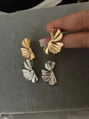 Guld- och silverfärgade örhängen - Två par örhängen i metall, ett par i guld och ett par i silver. Båda har en unik, veckad design som påminner om snäckskal eller solfjädrar. Perfekta för att ge en cool touch till din outfit.