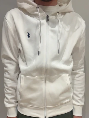 Vit hoodie med dragkedja från Polo Ralph Lauren - Snygg vit hoodie från Polo Ralph Lauren med dragkedja framtill, två fickor och justerbar huva med snören. Klassisk blå logga broderad på bröstet. Tillverkad i mjukt bomullsmaterial för en skön känsla.