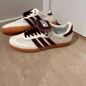 Adidas Wales Bonner sneakers vita/bruna - Snygga Adidas Wales Bonner sneakers i vitt och mörkbrunt med klassiska tre ränder på sidan och gummisula. Skorna har en unik detalj med kontrastsömmar på plösen och är tillverkade i en mix av mocka och textil. Perfekta för dig som gillar retrostil och streetwear.