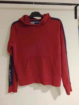 Röd hoodie från Gant RIKTIGT EFTERTRAKTAD🤌🏼 - Snygg röd hoodie från Gant med huva och känguruficka framtill. Marinblå ränder längs ärmarna med vit GANT-logga ger en sportig vibe. Tillverkad i mjukt material, perfekt för chill dagar. Klassisk passform och ribbade muddar vid ärmslut och nederkant. Mycket eftertraktad Gant tröja och är det mycket fint skick! FÖRST I KVARN👌🏼finns inga många av denna sort🤌🏼