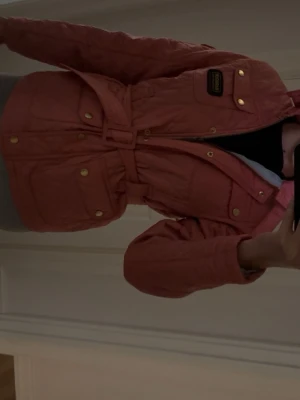 Rosa quiltad jacka från Barbour - Snygg rosa quiltad jacka från Barbour med bälte i midjan och guldiga knappar. Jackan har flera fickor framtill och ett Barbour-märke på bröstet. Perfekt för dig som vill ha en trendig och bekväm jacka med coola detaljer.