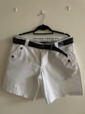 Klassiska vita shorts från Pelle P (L) - Klassiska vita shorts från Pelle P med originalskärp och stilrena nautiska detaljer. Tidlös design som passar perfekt till sommaren, segling, golf eller en avslappnad coastal look. Lätt och bekvämt material med dekorativa knappar och broderad logotypdetalj. Enkla att matcha med både stickat och skjortor.  Kan innehålla tecken på slitage.