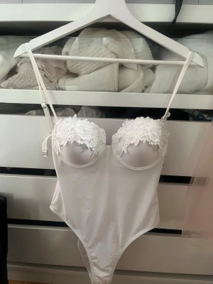 Vit spetsbody med tunna axelband - Snygg vit body med tunna axelband och vadderade kupor dekorerade med vacker spets. Bodyn har en slät, glansig yta och en feminin passform. Perfekt för dig som gillar romantiska detaljer och stilren design.