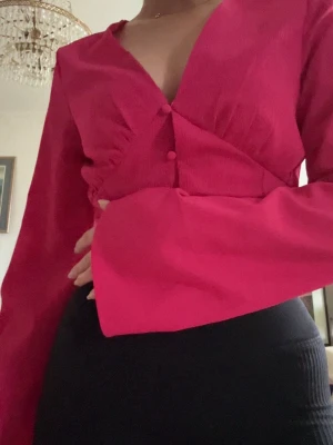 Rosa blus med vida ärmar - Trendig rosa blus med djup v-ringning och knappar framtill. Blusen har långa, vida ärmar och figurnära passform i midjan. Perfekt för dig som vill sticka ut med en färgstark och snygg look. Materialet är lätt och något strukturerat.
