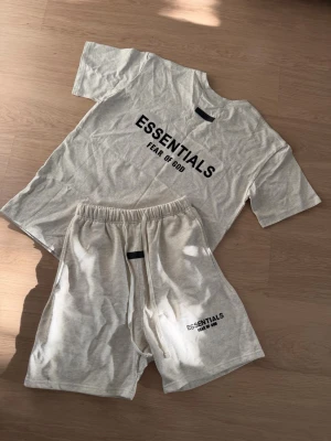 Essentials Fear of God loungewear set - Beige loungewear set från Essentials Fear of God med t-shirt och shorts. T-shirten har svart logotyptryck framtill och rund hals. Shortsen har resår i midjan och dragsko samt logotyptryck på benet. Perfekt för chill dagar hemma.