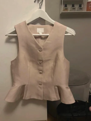 Beige väst med volangkant och knappar - Stilren beige väst med v-ringning och dekorativa knappar framtill. Västen har en snygg volangkant nedtill som ger en trendig touch. Perfekt att styla över en skjorta eller t-shirt för en cool look.
