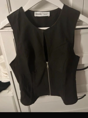 Svart väst med dragkedja från Bershka - Stilren svart väst från Bershka med rak passform och dragkedja framtill. Perfekt att styla över en t-shirt eller skjorta för en trendig look. Västen har en enkel design utan krage och är tillverkad i ett mjukt material.