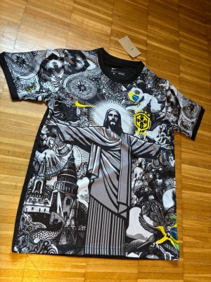  Brasilien fotbollströja, jesus grafisk t-shirt - Unik t-shirt från Nike med Brasil-tema och grafiskt tryck av Kristusstatyn och brasilianska symboler. Svart och grå bas med inslag av gult och blått. Rund hals och korta ärmar. Perfekt för dig som vill sticka ut med en cool och konstnärlig look. Jag fick de som present men storleken passar inte mig.