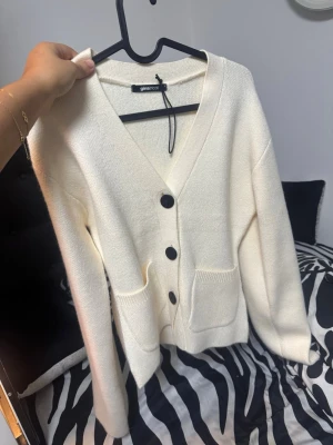 Vit kofta från Gina Tricot - Stilren vit kofta från Gina Tricot med djup V-ringning och stora svarta knappar. Koftan har två praktiska fickor framtill och är tillverkad i ett mjukt stickat material. Perfekt för lager-på-lager och enkel att matcha.