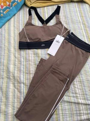 Brunt set från Alo med sport-bh och tights - Snyggt tvådelat set från Alo i brun nyans med svarta och vita detaljer. Setet består av en sport-bh med korsade axelband och matchande tights, båda med elastisk midja och vit piping. Perfekt för en trendig och sportig look.