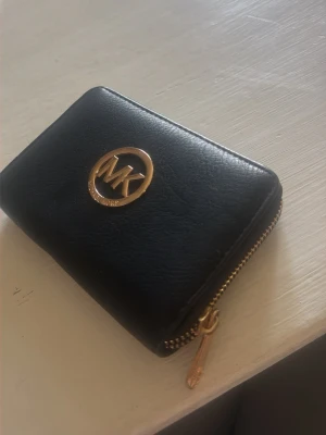 Svart Michael Kors plånbok i läder - Snygg svart plånbok från Michael Kors i äkta läder med guldfärgade detaljer och dragkedja runtom. Plånboken har flera kortfack och ett myntfack med dragkedja. Perfekt storlek för att få plats med det viktigaste och enkel att ta med sig.