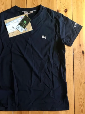 Marinblå Burberry t-shirt i bomull - Snygg marinblå t-shirt från Burberry med klassisk rund halsringning och korta ärmar. Tillverkad i mjuk bomull och har Burberrys ikoniska logga broderad på bröstet. Perfekt basplagg för en clean och stilren look.