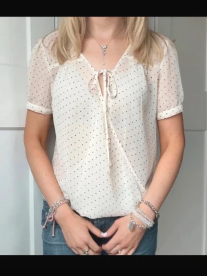 Prickig blus med knytning i halsen - Söt ljusbeige blus med små svarta prickar och knytning i halsen. Blusen har korta ärmar och är tillverkad i ett tunt, lätt och transparent material. Mindre / tajt vid armarna annars jättesöt mellan S och M skulle jag säga i storlek. Köpt för 399, ny skick