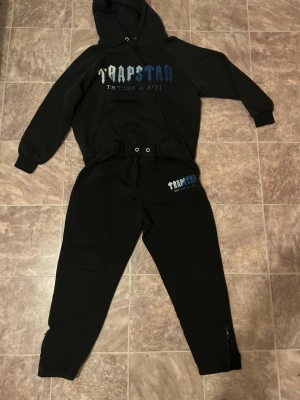 Trapstar svart hoodie och byxset - Säljer ett svart set från Trapstar med hoodie och matchande byxor. Hoodien har tryckt logga i blått och vitt framtill och byxorna har logga på ena benet. Setet har ribbade muddar och byxorna har dragkedja vid benslut. Perfekt för en avslappnad stil.