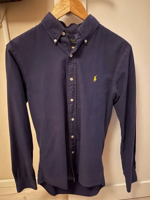 Skjorta från Polo Ralph Lauren - Klassisk mörkblå skjorta från Polo Ralph Lauren med button-down krage och den ikoniska gula loggan broderad på bröstet. Skjortan har långa ärmar, vita knappar och är tillverkad i mjuk bomull. Perfekt för en clean och stilren look.