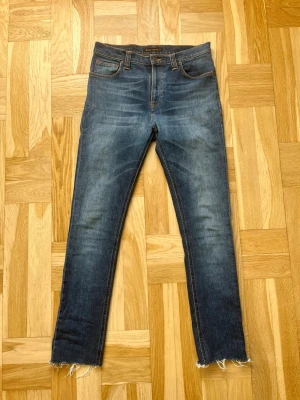 Blå skinny jeans från Nudie Jeans archive fashion - Säljer ett par blå skinny jeans från Nudie Jeans med klassisk femficksdesign och råa, fransiga benslut. Jeansen har snygga slitningar och kontrastsömmar. Perfekta för en avslappnad och trendig look.