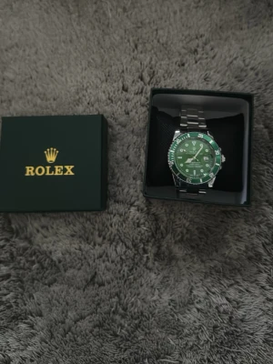 klocka med grön urtavla - Stilren klocka med grön urtavla och silverfärgad länk i metall. Klockan har tydliga index, datumvisning och klassisk rund form. Levereras i originalask med kudde. Perfekt accessoar för dig som vill sticka ut med exklusiv känsla.