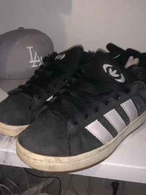 Adidas Campus svarta sneakers - Svarta Adidas Campus sneakers med klassiska vita ränder på sidorna och vit sula. Skorna har svart mockaöverdel, svarta skosnören och Adidas logga på plösen. Perfekta för en streetwear-look och passar till det mesta.
