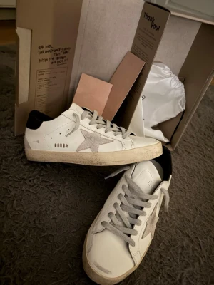 Golden Goose vita sneakers med stjärna - Golden Goose sneakers i vitt läder med grå mockadetaljer och ikonisk stjärna på sidan. Skorna har grå skosnören, svart hälparti och en vintage-inspirerad sula. Perfekta för dig som gillar en avslappnad och trendig stil.