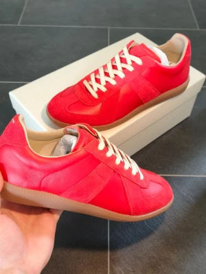 Röda sneakers från Maison Margiela German Army Trainer GATS - Snygga röda sneakers från Maison Margiela med ovandel i både mocka och läder. Skorna har vita snören, beige innersula och en klassisk gummisula. Perfekt för dig som vill sticka ut med stilrena och trendiga sneakers.