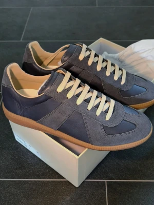 Blå Navy GATS Maison Margiela German Army Trainer - Snygga blå sneakers från Maison Margiela med ovandel i mocka och läder, vita snören och klassisk gummisula. Skorna har en stilren och tidlös design som passar perfekt till streetwear och casual outfits.