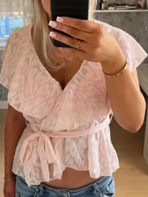 Ljusrosa omlottblus med knytband - Söt ljusrosa omlottblus med diskret mönster och volangig krage. Blusen har korta ärmar och knyts i midjan med ett matchande band. Perfekt för en trendig och feminin look.
