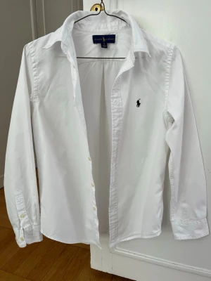 Ralph Lauren skjorta - Klassisk vit skjorta från Ralph Lauren med lång ärm och knappar framtill. Skjortan har en stilren design med mörkblå, broderad logga på bröstet och är tillverkad i bomull. Skicket är väldigt bra och storleken är 16! Hör av dig vid minsta fundering!