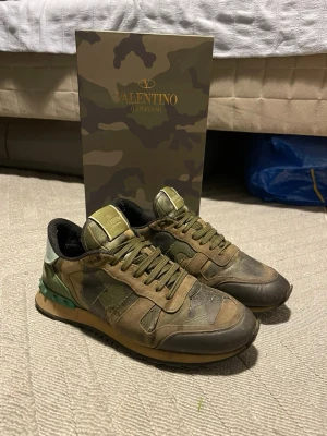 Valentino Garavani camo sneakers - Snygga sneakers från Valentino Garavani i camomönster med gröna, bruna och svarta toner. Skorna har klassisk låg siluett, svarta detaljer och nitar på hälen. Tillverkade i en mix av läder och textil för en exklusiv känsla.