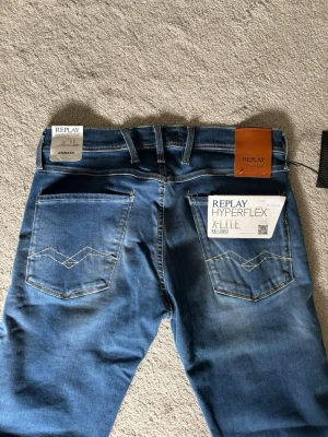 Replay Anbass Blå - Säljer ett par oanvända Replay Anbass Hyperflex jeans i blå tvätt med snygga kontrastsömmar. Modellen har raka ben, klassiska bakfickor och en brun Replay-lapp i midjan. Materialet är stretchigt för extra komfort och rörlighet. Slim passform, storlek 31/34.